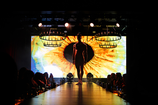 A performer walks down a runway with a vibrant, artistic backdrop while an audience watches from their tables.