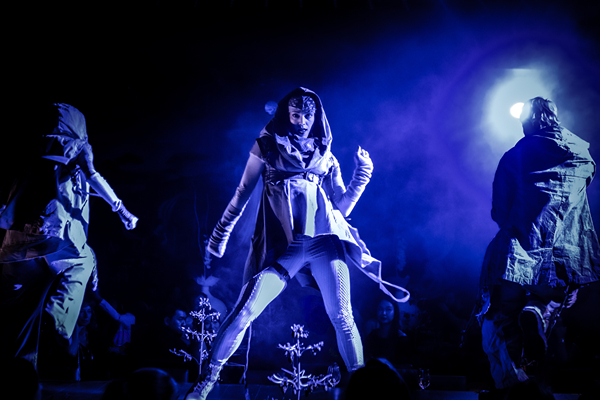 Three performers in dramatic costumes engage in a theatrical dance under blue lighting, creating an immersive dining experience.