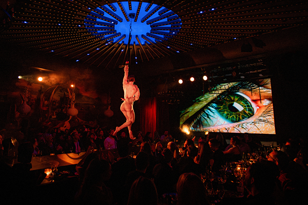 A performer hangs from a chain above an audience in a vibrant dining theater with colorful lights and a large eye projection.