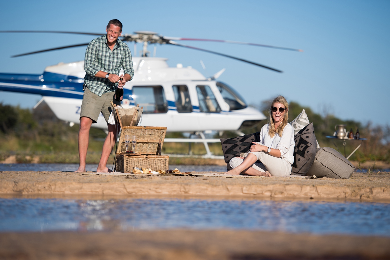 A couple celebrates with champagne on a sandy beach, a helicopter in the background, set for a romantic honeymoon escape.