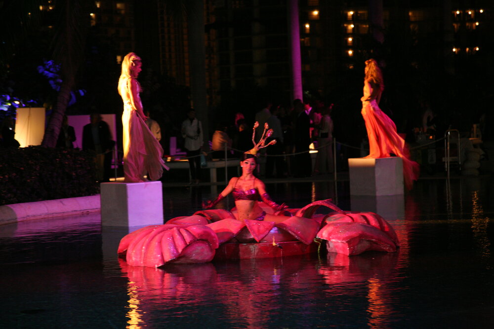 A performer in a pink costume sits on a large lotus flower in a pool, flanked by two models in flowing garments.