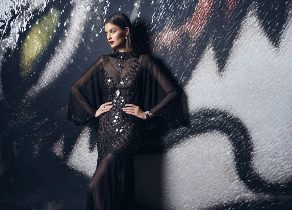 Model in an elegant, dark gown with sequins poses against a dramatic painted backdrop, showcasing fashion artistry.