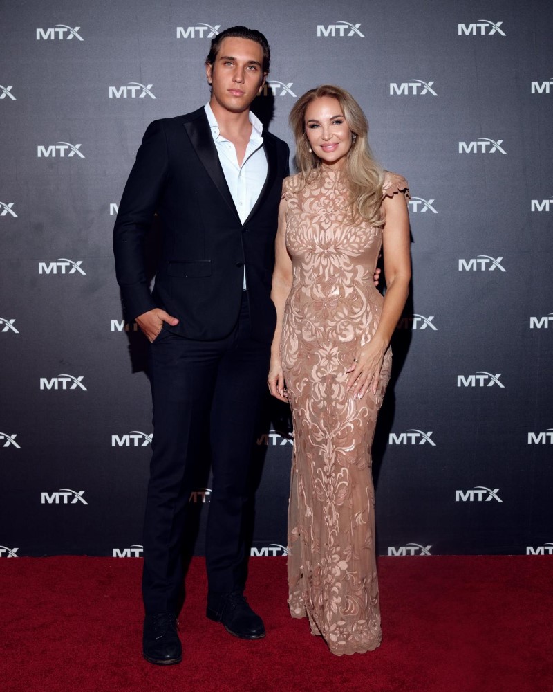Ema Savahl and a male companion pose together on a red carpet, showcasing elegant fashion against a branded backdrop.