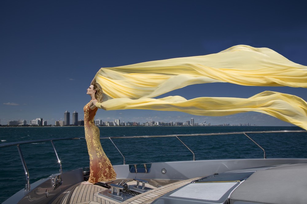 Model in a flowing yellow fabric stands on a yacht, overlooking a city skyline against a clear blue sky.