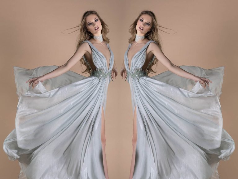 Model in a flowing light blue gown with intricate details, embodying grace and elegance in a soft pastel backdrop.