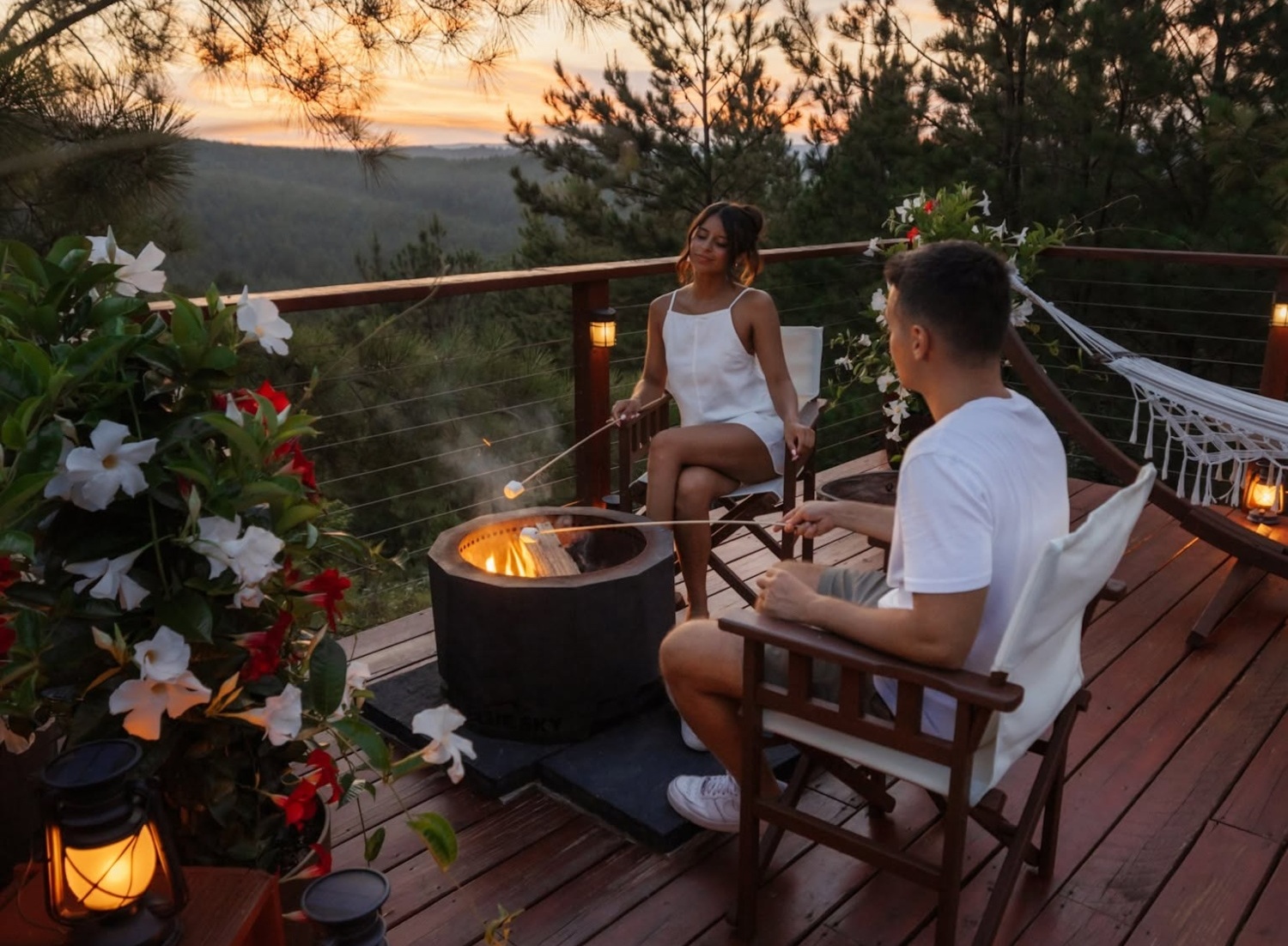 Couple roasting marshmallows by a fire pit on a balcony surrounded by greenery at sunset.