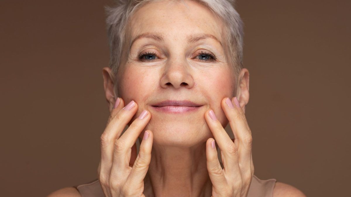 Mature woman applying illuminating primer makeup with a gentle smile and a soft touch to her face.