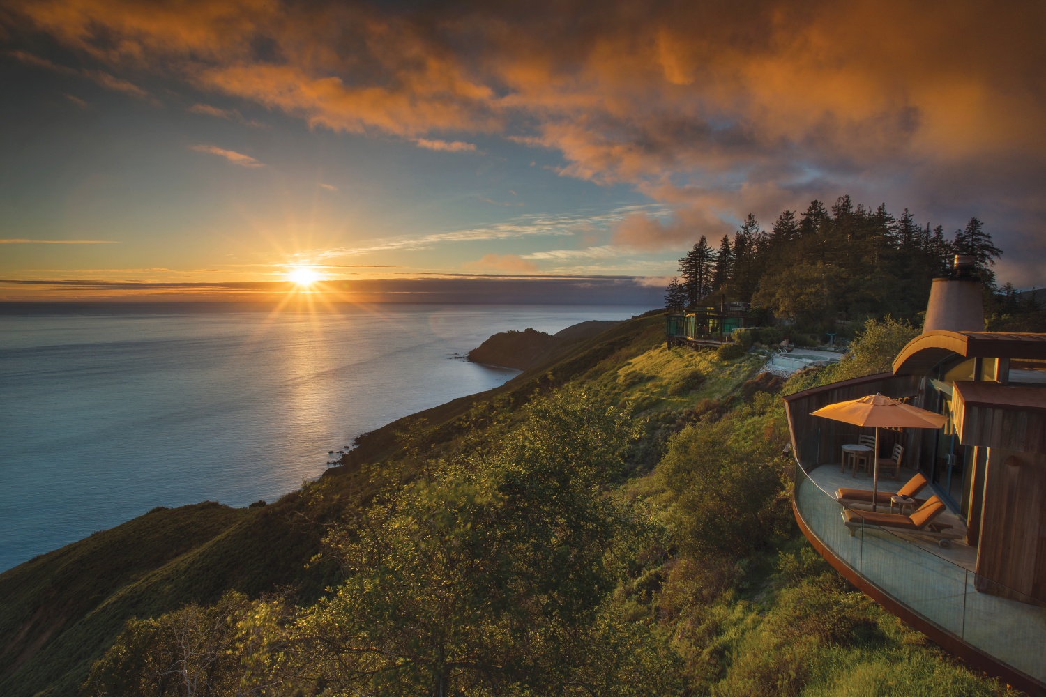 Sunset over a coastal landscape, highlighting lush hills and a modern building with a deck and umbrella.