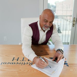 Man signing documents at a wooden table with a retirement plan logo, focusing on financial details and planning.