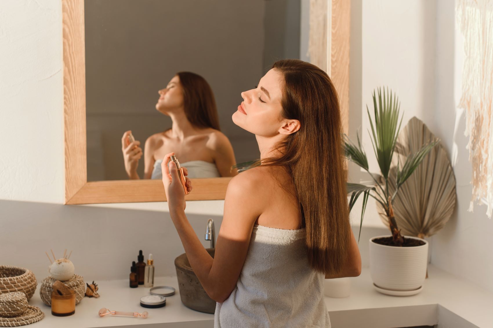 Woman applying perfume in front of a mirror, showcasing a bathroom setting with beauty products and a plant.