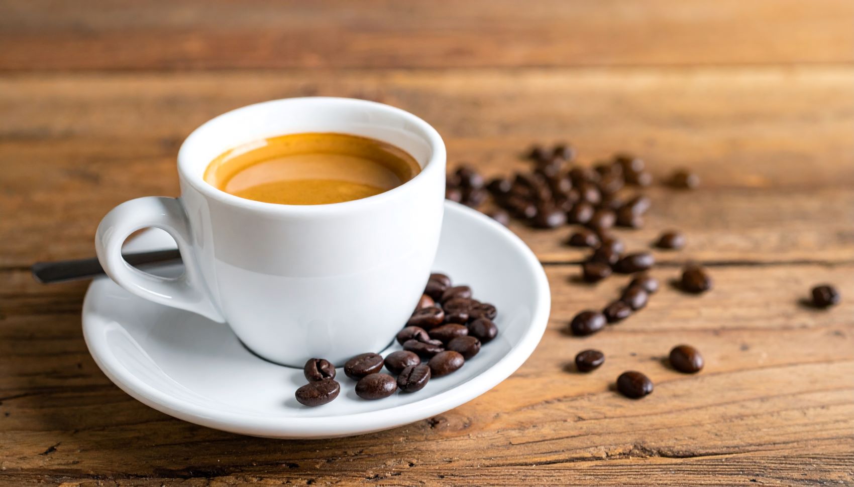 A steaming cup of coffee on a saucer surrounded by scattered coffee beans on a wooden table.