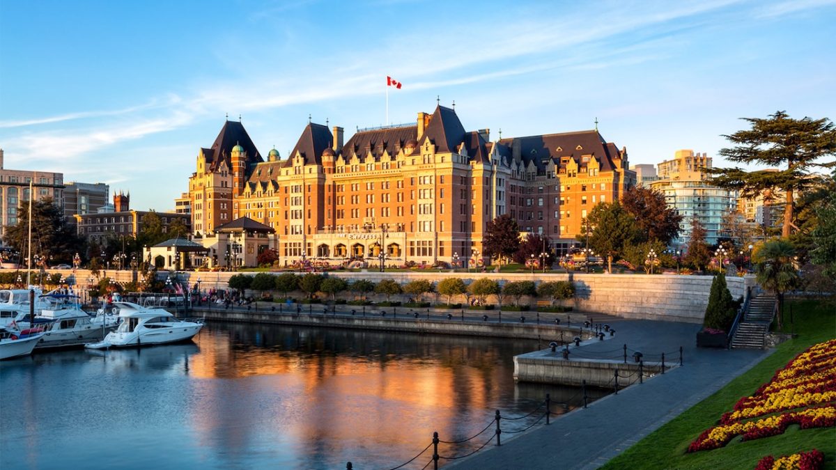 Fairmont Empress hotel reflecting in the water at sunset, with boats in the foreground and colorful flowerbeds along the shore.