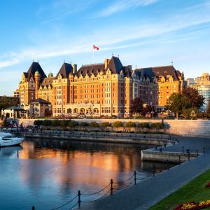 Fairmont Empress hotel reflecting in the water at sunset, with boats in the foreground and colorful flowerbeds along the shore.