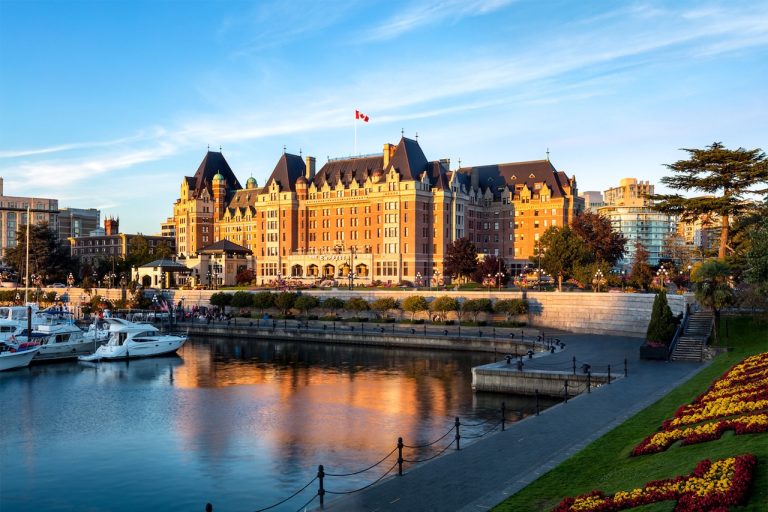 Fairmont Empress hotel reflecting in the water at sunset, with boats in the foreground and colorful flowerbeds along the shore.
