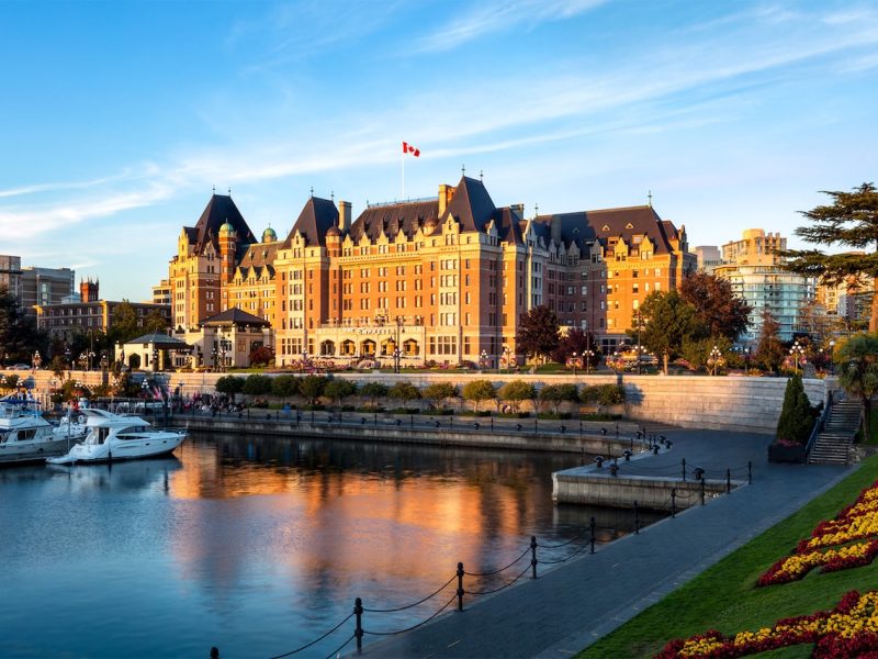 Fairmont Empress hotel reflecting in the water at sunset, with boats in the foreground and colorful flowerbeds along the shore.