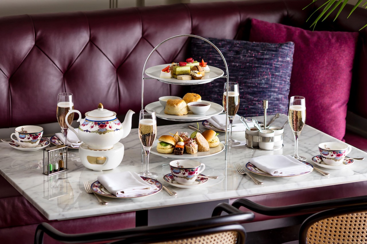 Elegant afternoon tea setup featuring a floral teapot, fine china, scones, sandwiches, and sparkling wine in a luxurious setting.