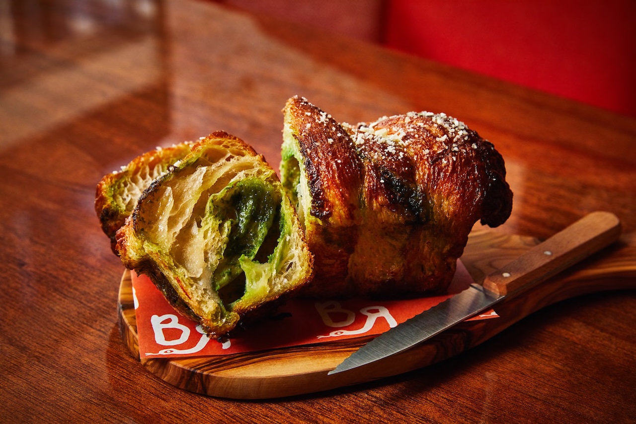 A sliced green bread roll with a crispy crust on a wooden board, served with a knife on a colorful paper.