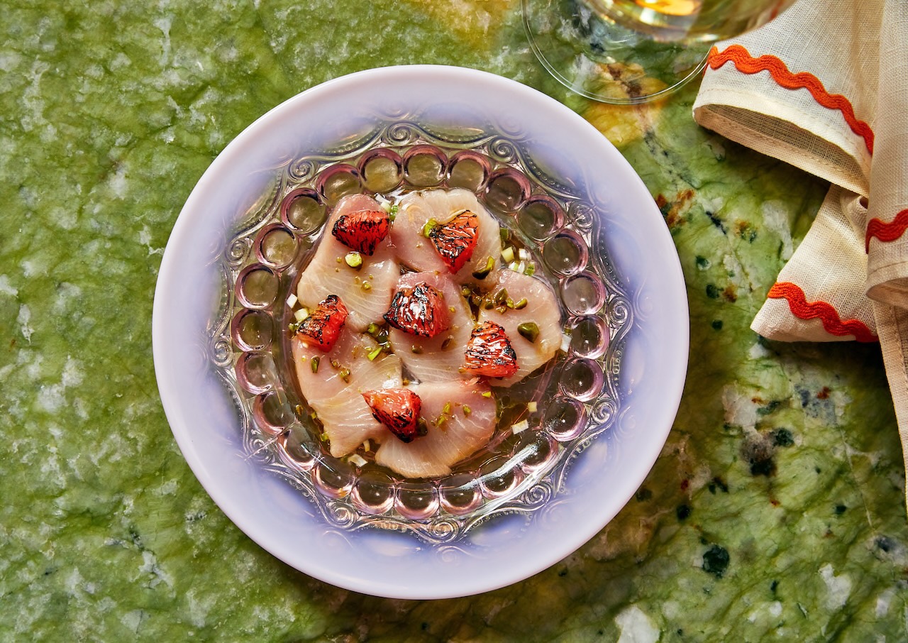 Sashimi-style fish garnished with charred strawberries and pistachios on a decorative plate, set on a green marble surface.