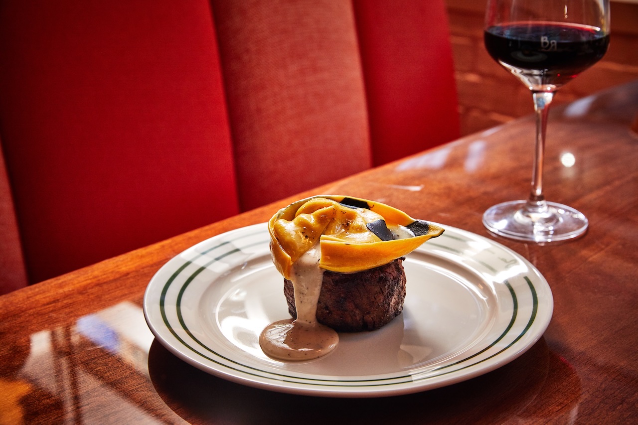 Steak topped with a creamy sauce and yellow ribbon-like pasta, presented on a white plate with green accents beside a glass of red wine.