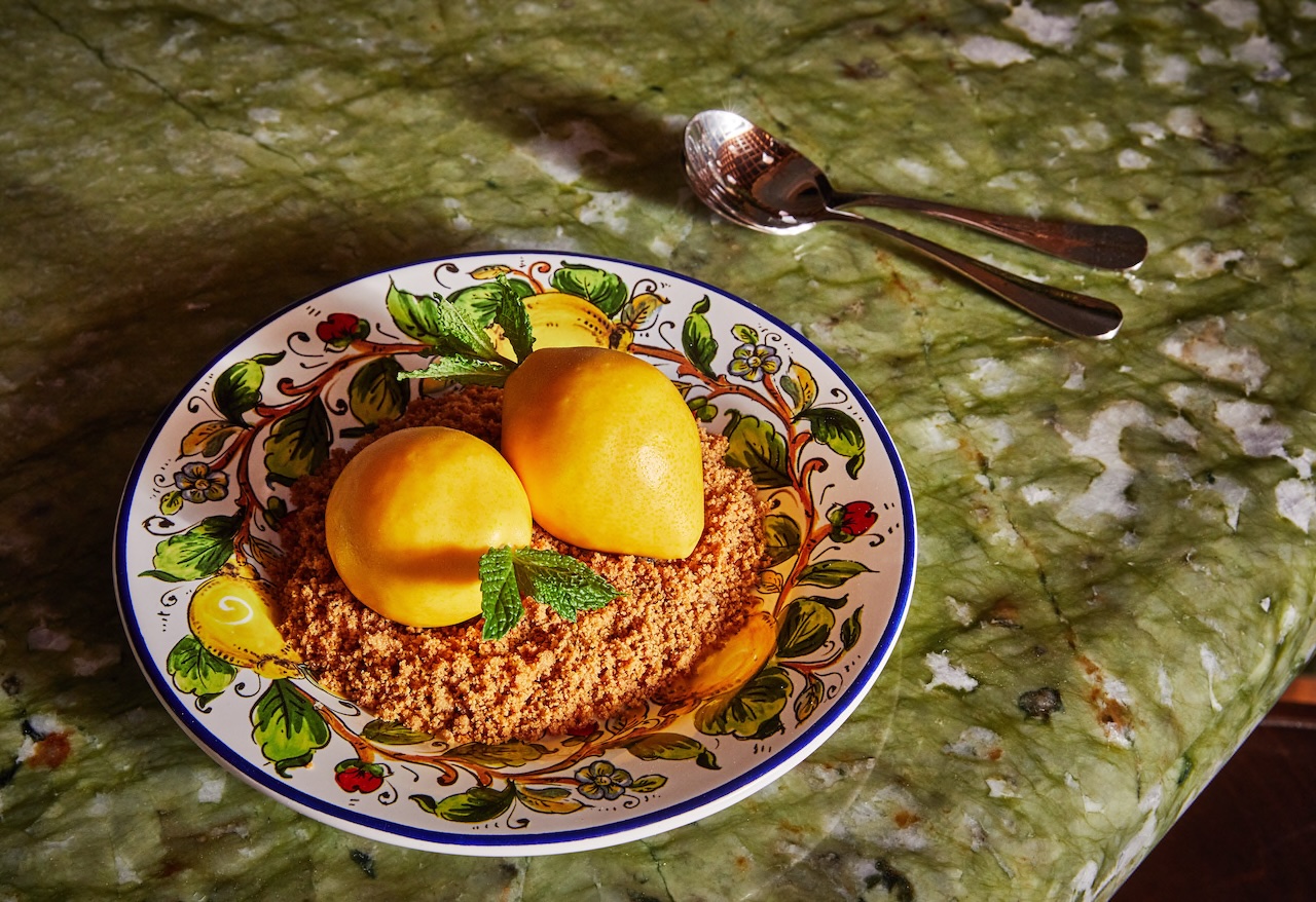 A decorative plate with two yellow lemons atop a layer of crumbly dessert, garnished with mint leaves on a green marble table.