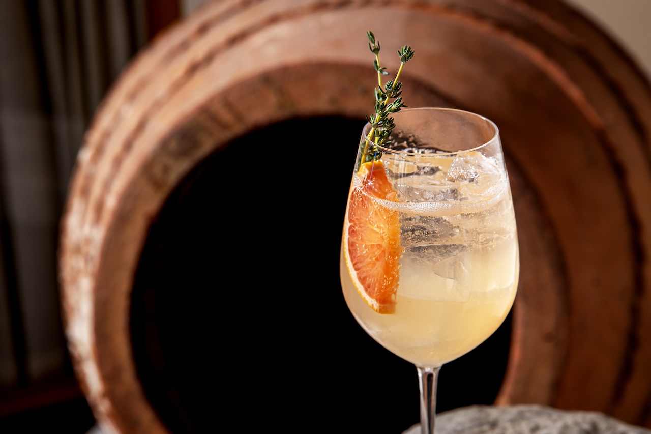 A refreshing cocktail garnished with an orange slice and thyme, set against rustic wooden barrels.