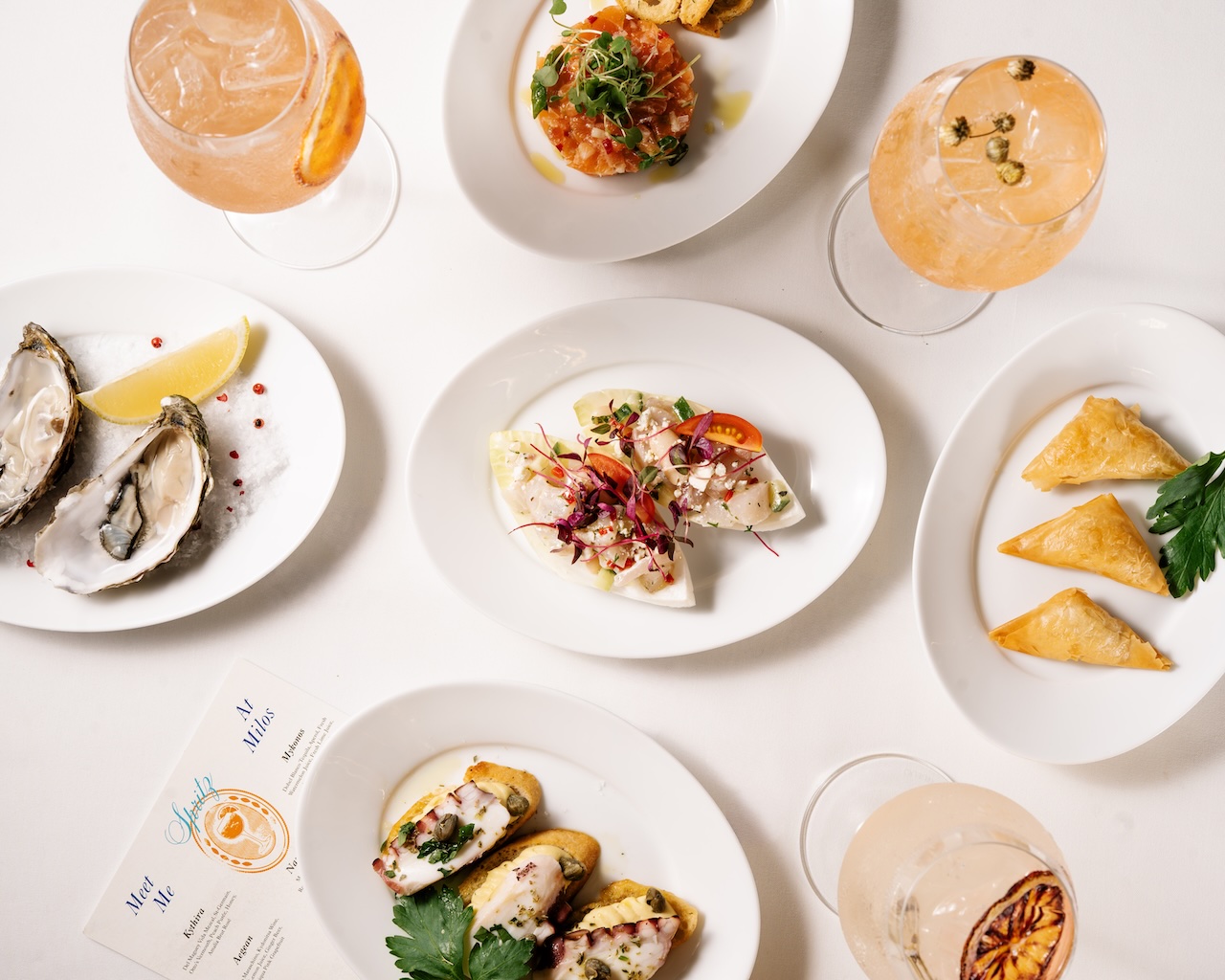 Colorful dishes including oysters, seafood salads, and pastries, accompanied by refreshing cocktails on a white tablecloth.