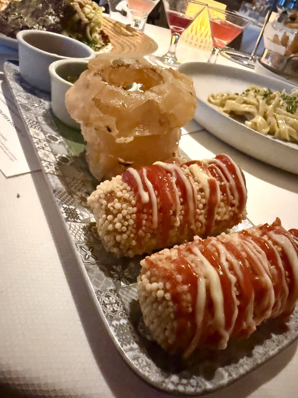 Hot dogs drizzled with sauce on a decorative plate, accompanied by various dipping sauces and elegant cocktails in the background.