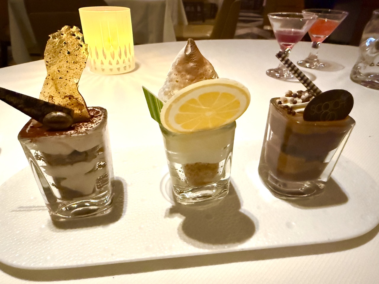 Three gourmet desserts in glass cups, featuring unique toppings, presented on a white plate with cocktails in the background.