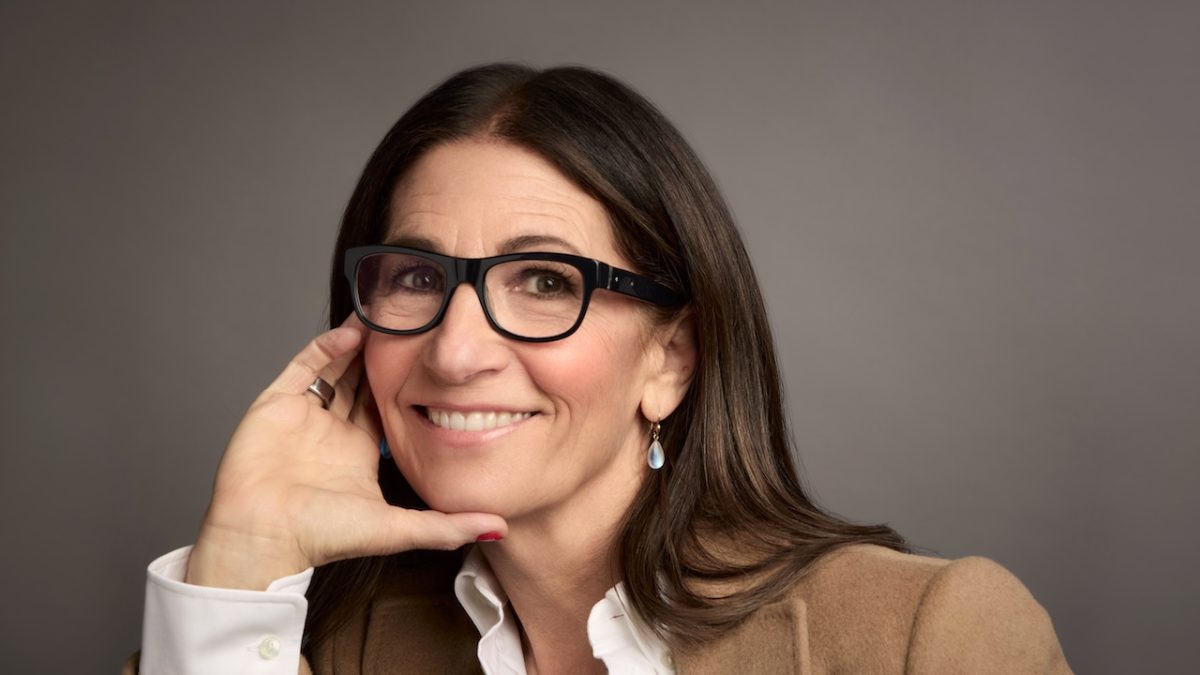 Bobbi Brown smiles while resting her hand on her face, wearing glasses and a beige jacket, promoting Jones Road Beauty.