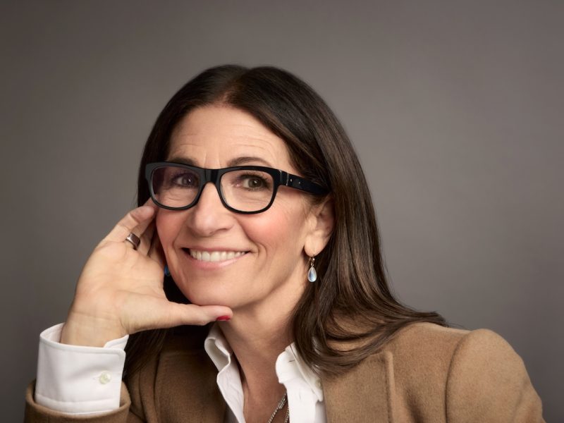 Bobbi Brown smiles while resting her hand on her face, wearing glasses and a beige jacket, promoting Jones Road Beauty.