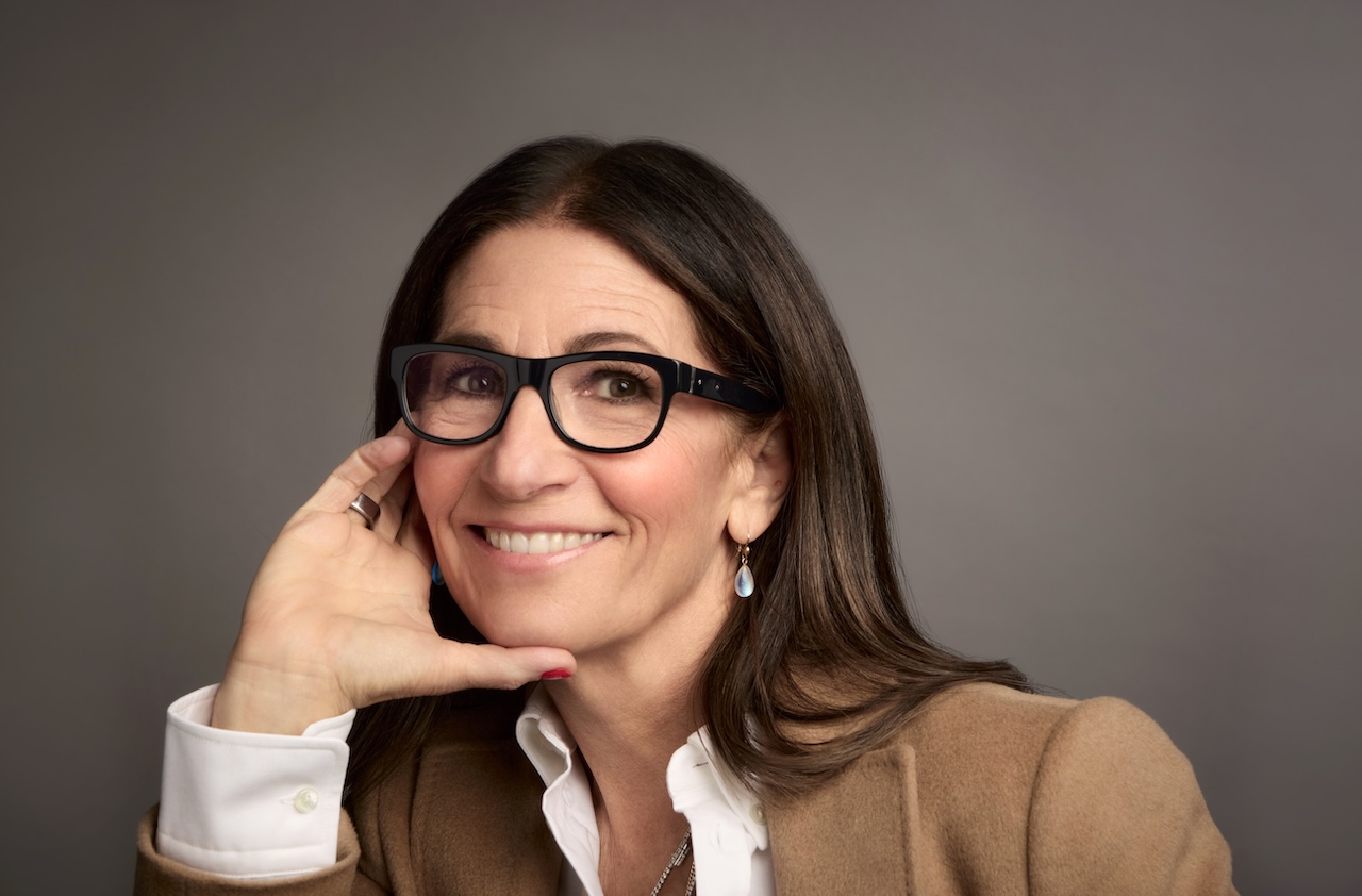 Bobbi Brown smiles while resting her hand on her face, wearing glasses and a beige jacket, promoting Jones Road Beauty.