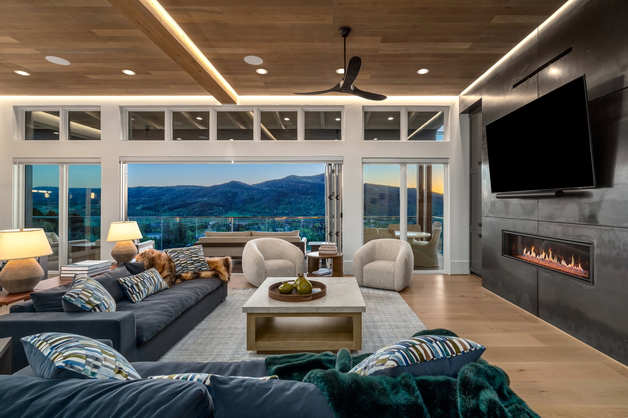 Modern living room featuring a stylish sofa, cozy chairs, and expansive windows showcasing mountain views at sunset.