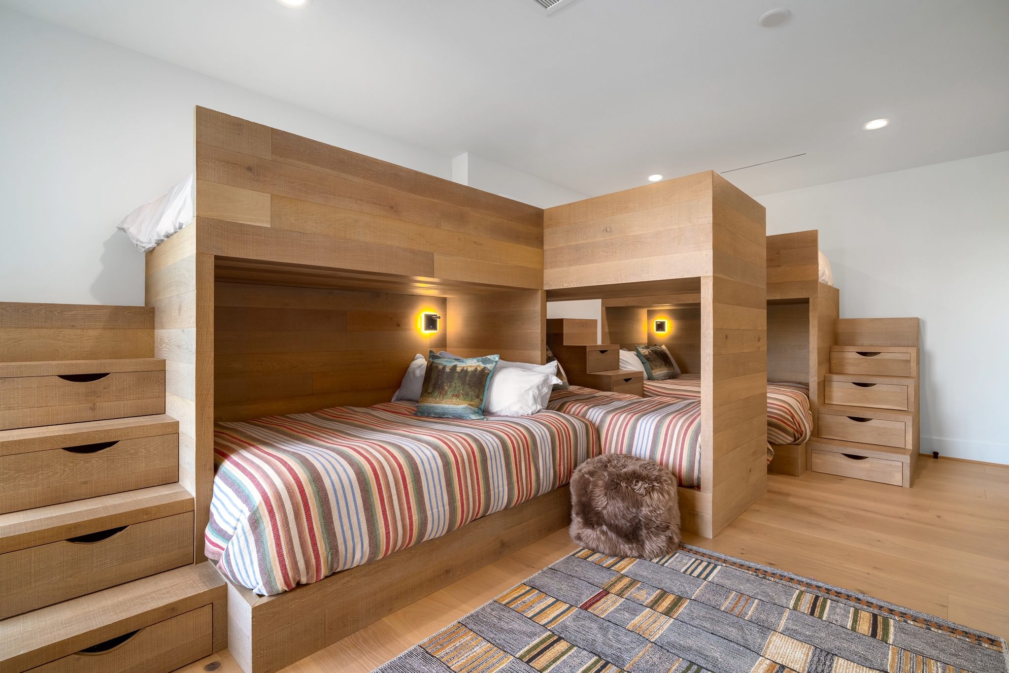 Bunk beds with wooden frames and striped bedding in a cozy room, featuring soft lighting and a textured rug.