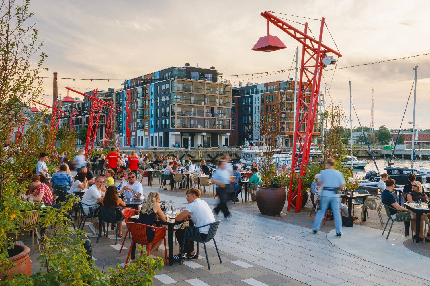 Crowded waterfront dining area with people enjoying meals, colorful buildings, and red cranes in the background near a marina.