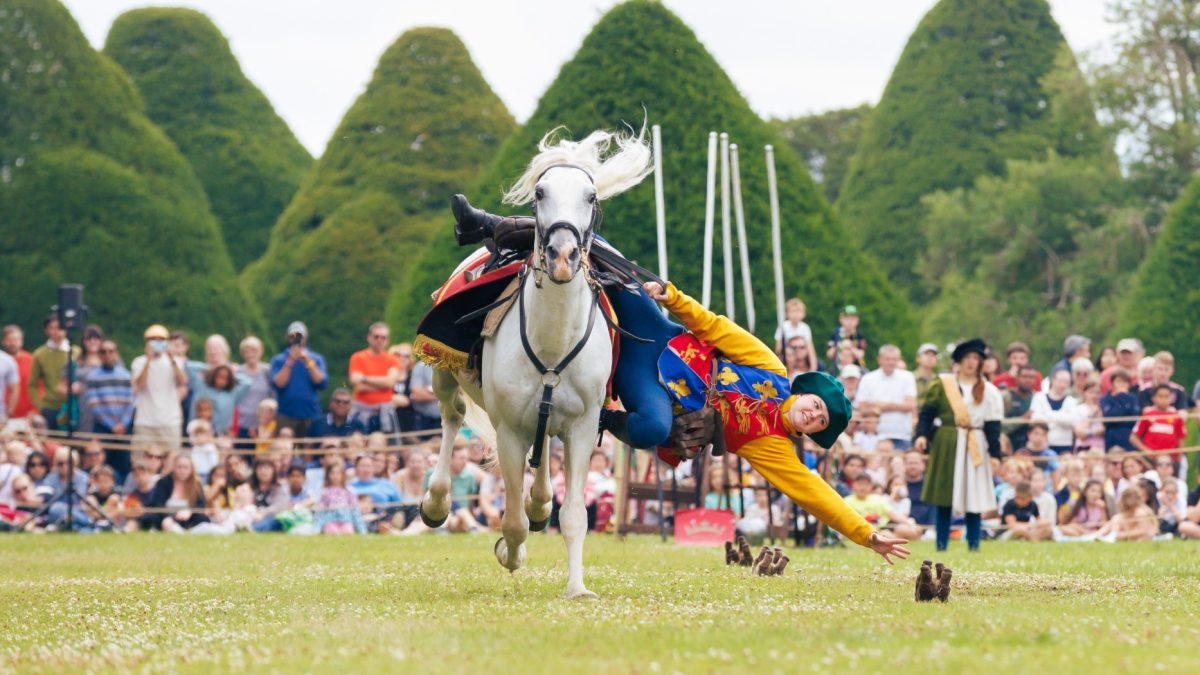 A performer in colorful costume falls from a galloping horse during a live show in front of a large audience at Hampton Court Palace.