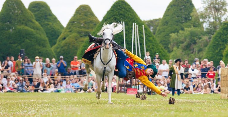 A performer in colorful costume falls from a galloping horse during a live show in front of a large audience at Hampton Court Palace.