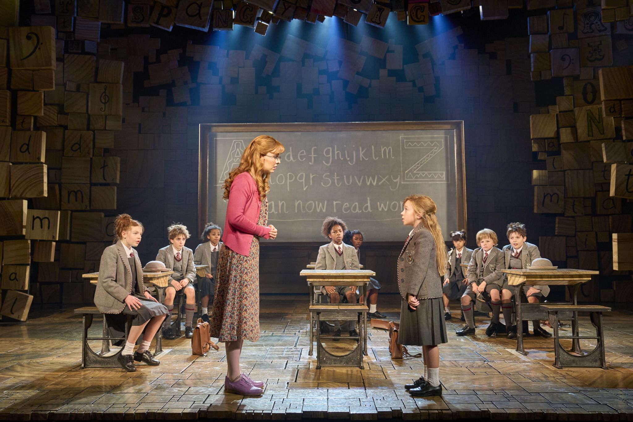 Scene from Matilda the Musical featuring characters in a classroom setting, with students seated at desks and two girls standing.