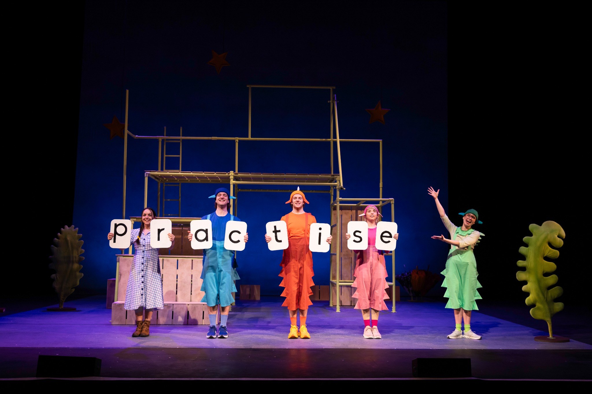 Five performers in colorful costumes hold letters spelling "practise" on stage, set against a bright blue background.