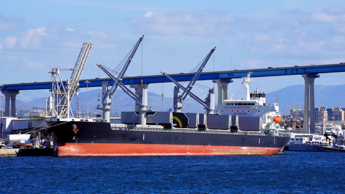 Cargo ship docked at a port, with cranes and industrial infrastructure visible in the background.