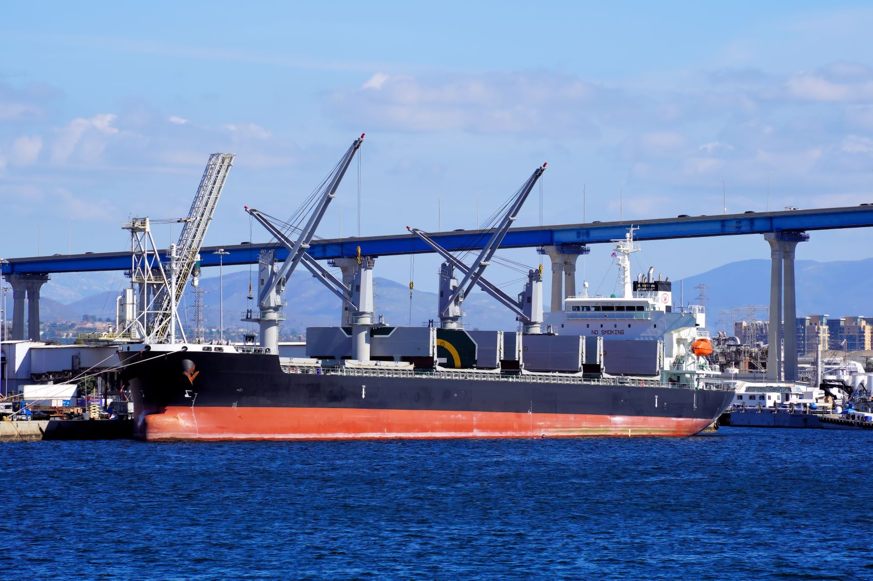 Cargo ship docked at a port, with cranes and industrial infrastructure visible in the background.