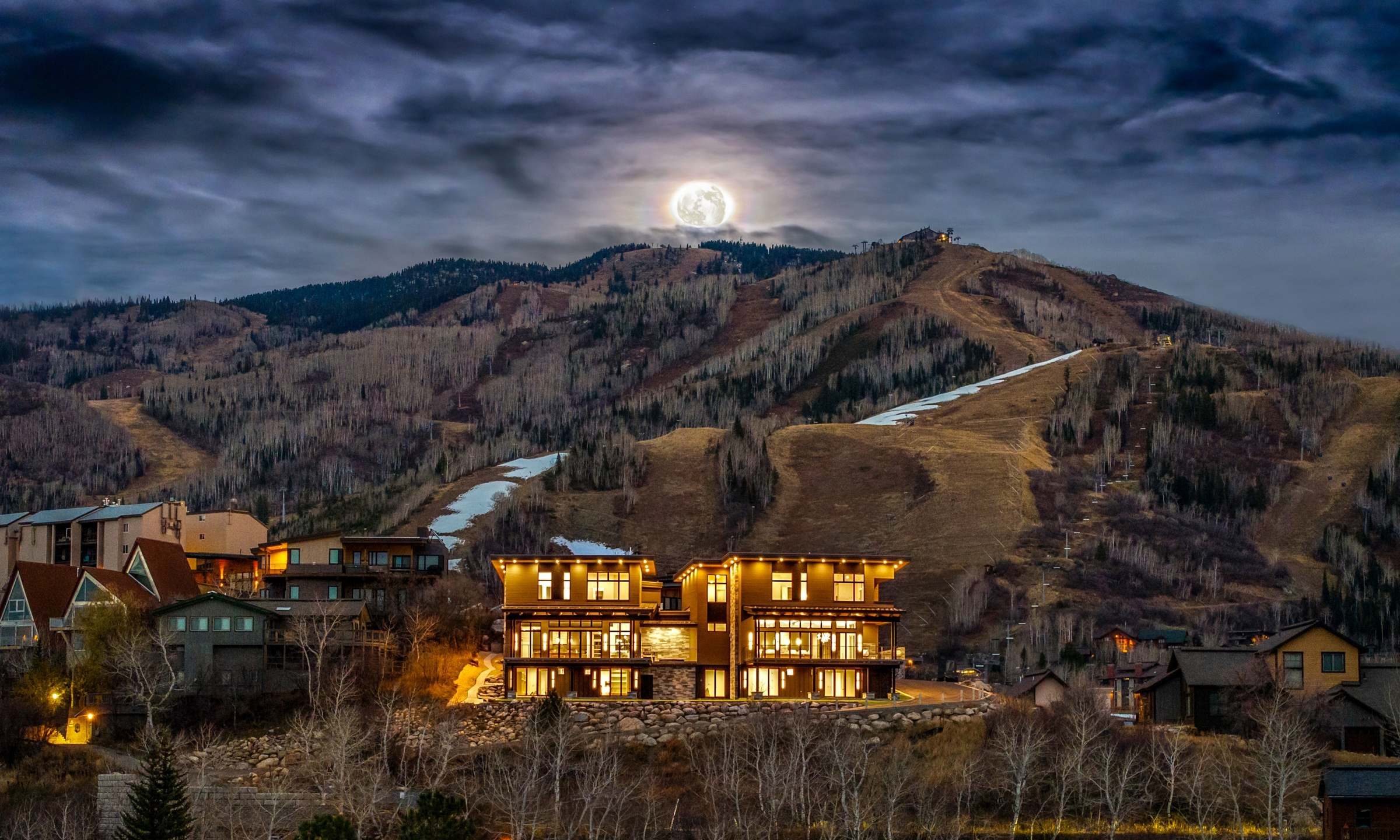 A stunning luxury home illuminated at night with a full moon rising above a mountain landscape in the background.