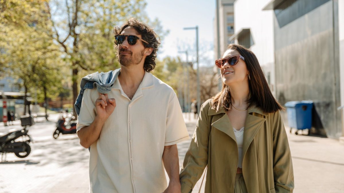 A couple walks hand in hand, smiling, wearing sunglasses, in a sunlit urban environment surrounded by trees.