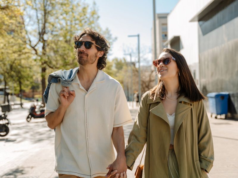 A couple walks hand in hand, smiling, wearing sunglasses, in a sunlit urban environment surrounded by trees.