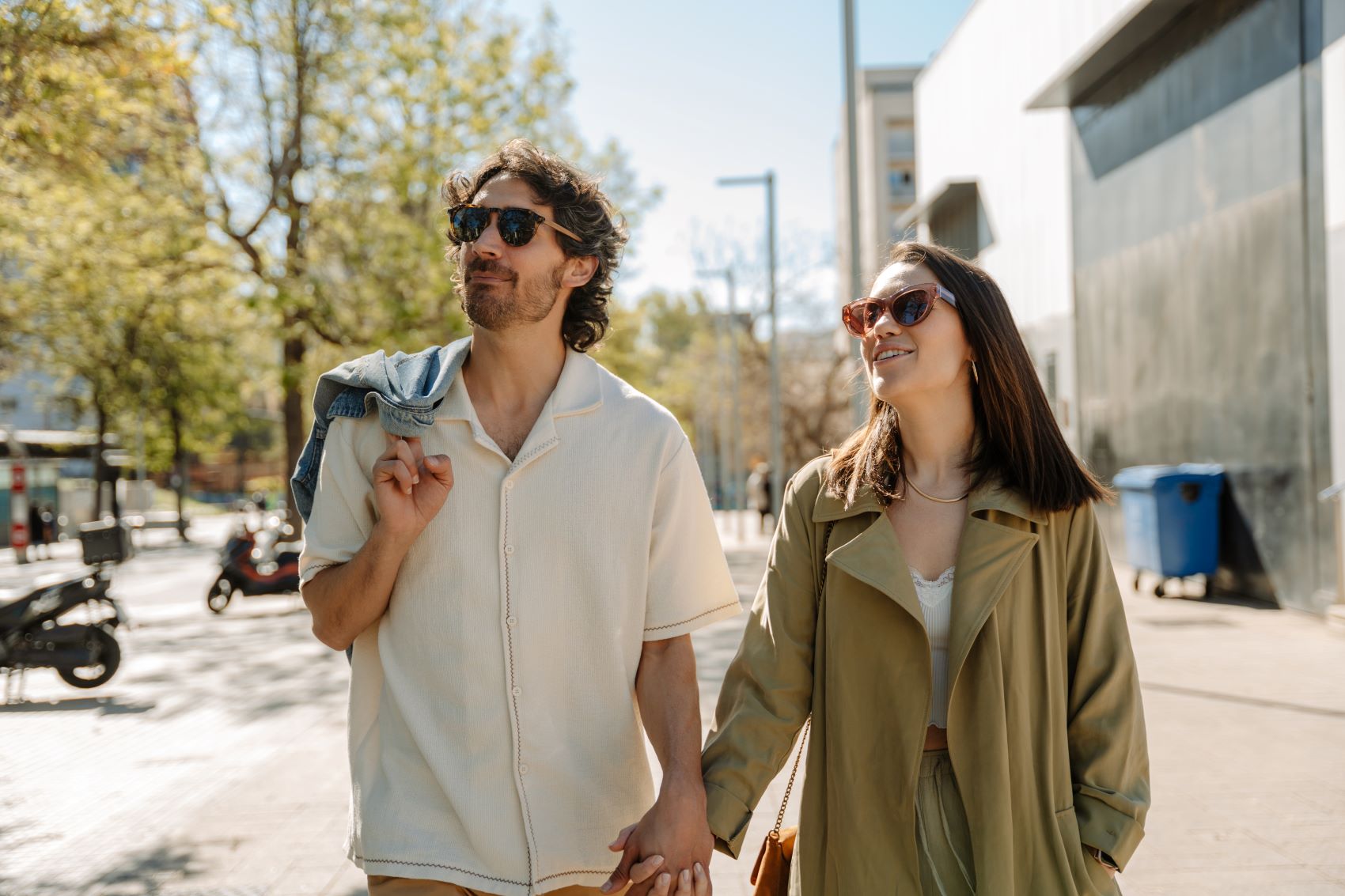 A couple walks hand in hand, smiling, wearing sunglasses, in a sunlit urban environment surrounded by trees.