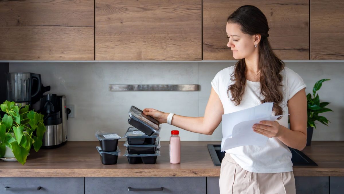 A woman checks meal prep containers and holds a document in a modern kitchen with plants and kitchen appliances.