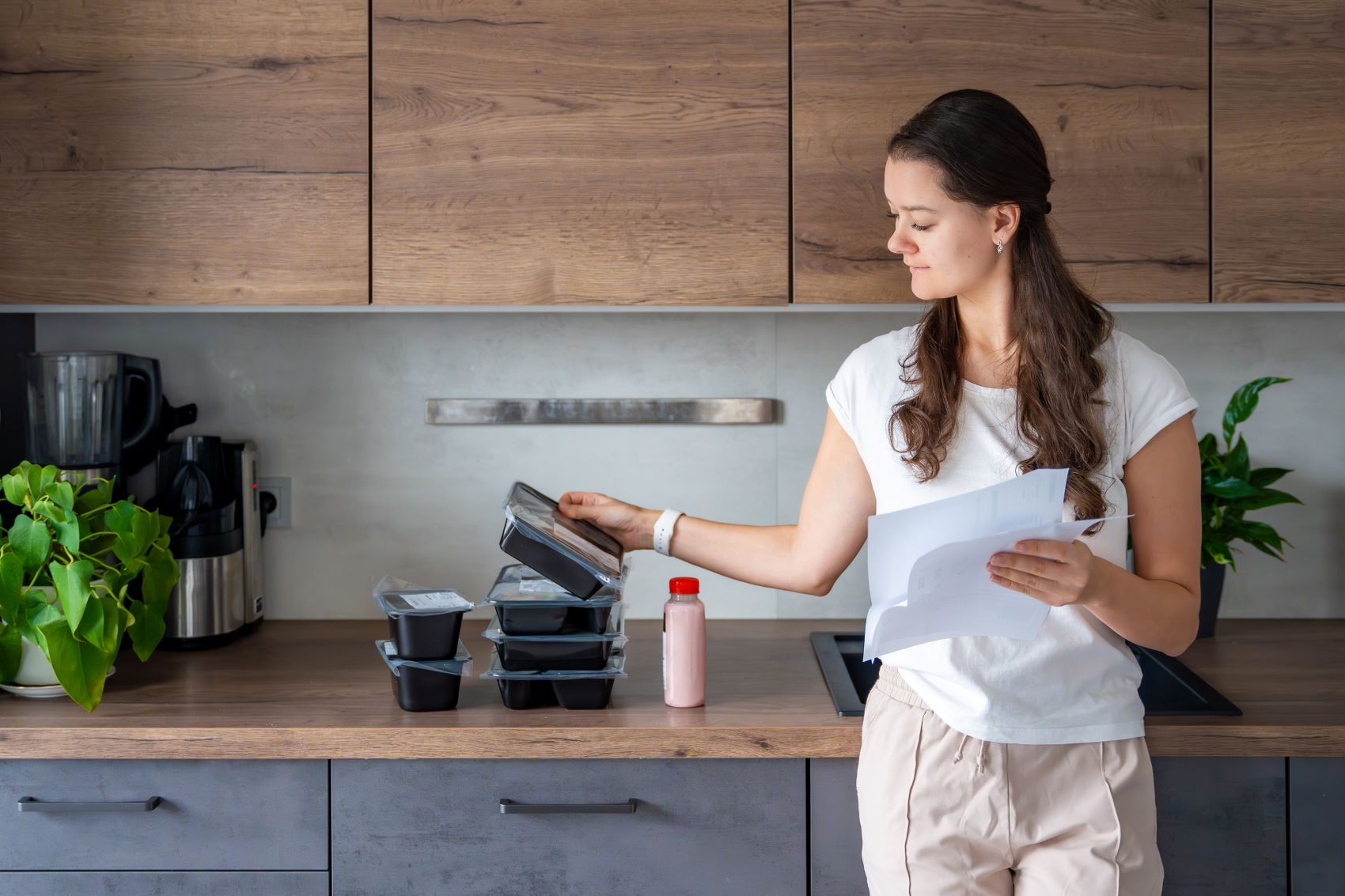 A woman checks meal prep containers and holds a document in a modern kitchen with plants and kitchen appliances.
