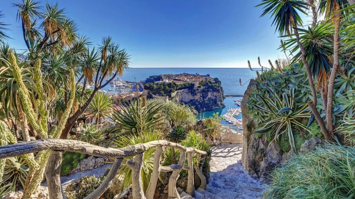 Lush greenery surrounds a stone staircase leading down to the Mediterranean coast with a view of a coastal town.