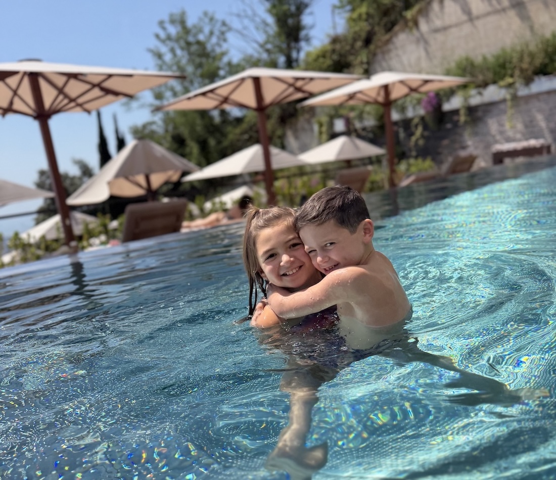 Two children smiling and hugging in a swimming pool, surrounded by umbrellas and a sunny outdoor setting.