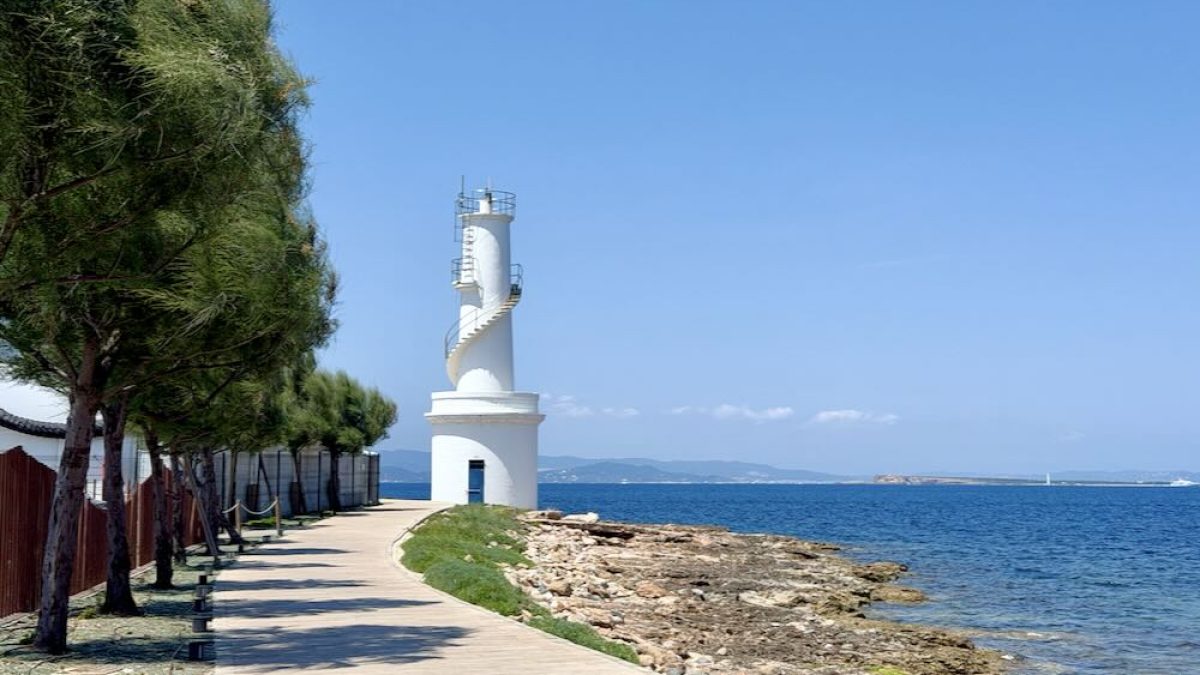 A coastal pathway in Formentera lined with trees, leading to a white lighthouse beside the turquoise sea under a clear blue sky.