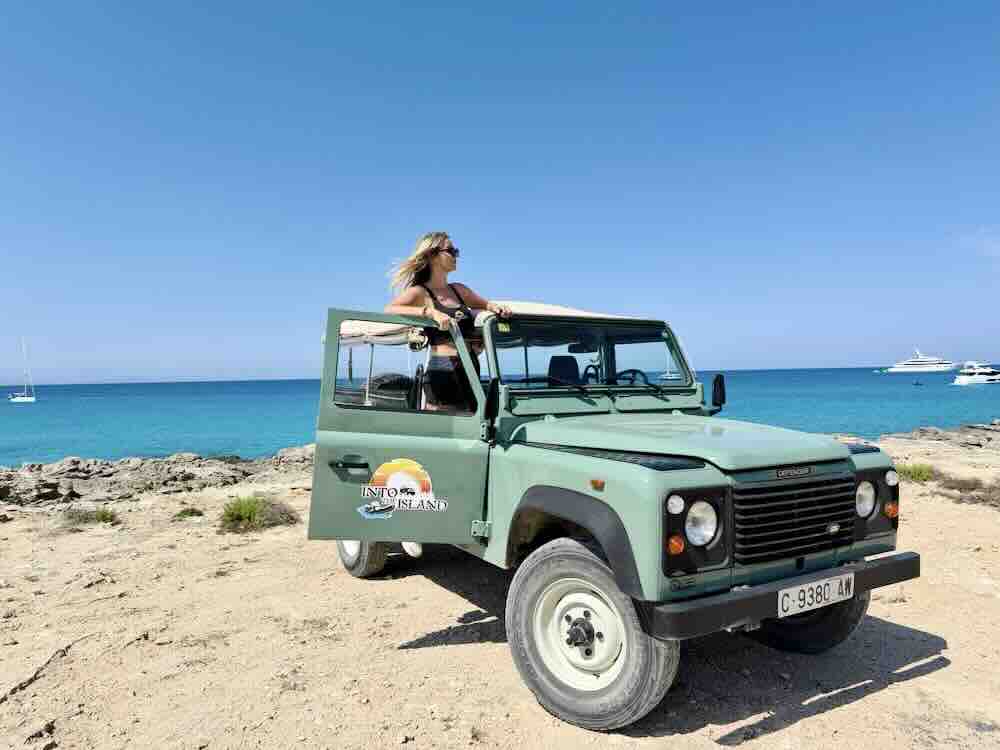 A woman enjoys the view from a green off-road vehicle near the beach on Formentera island, with boats in the background.